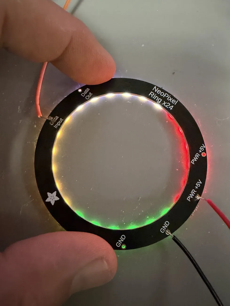 Close-up of the NeoPixel ring PCB held in hand, showing the solder joints on the pads and the ring glowing with gem colors