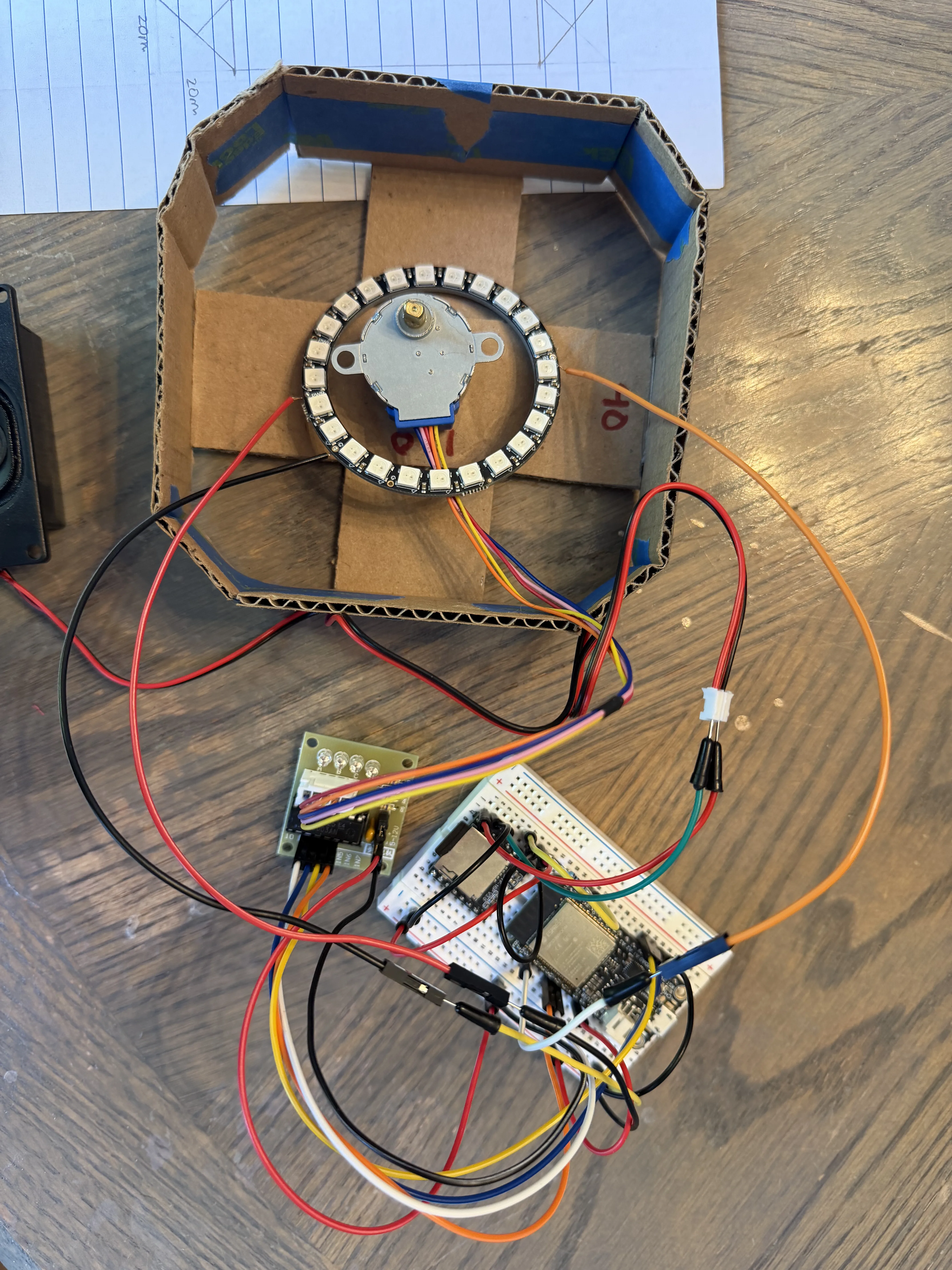 NeoPixel ring mounted directly on the stepper motor shaft, sitting inside the cardboard mockup housing with the full electronics stack wired in around it