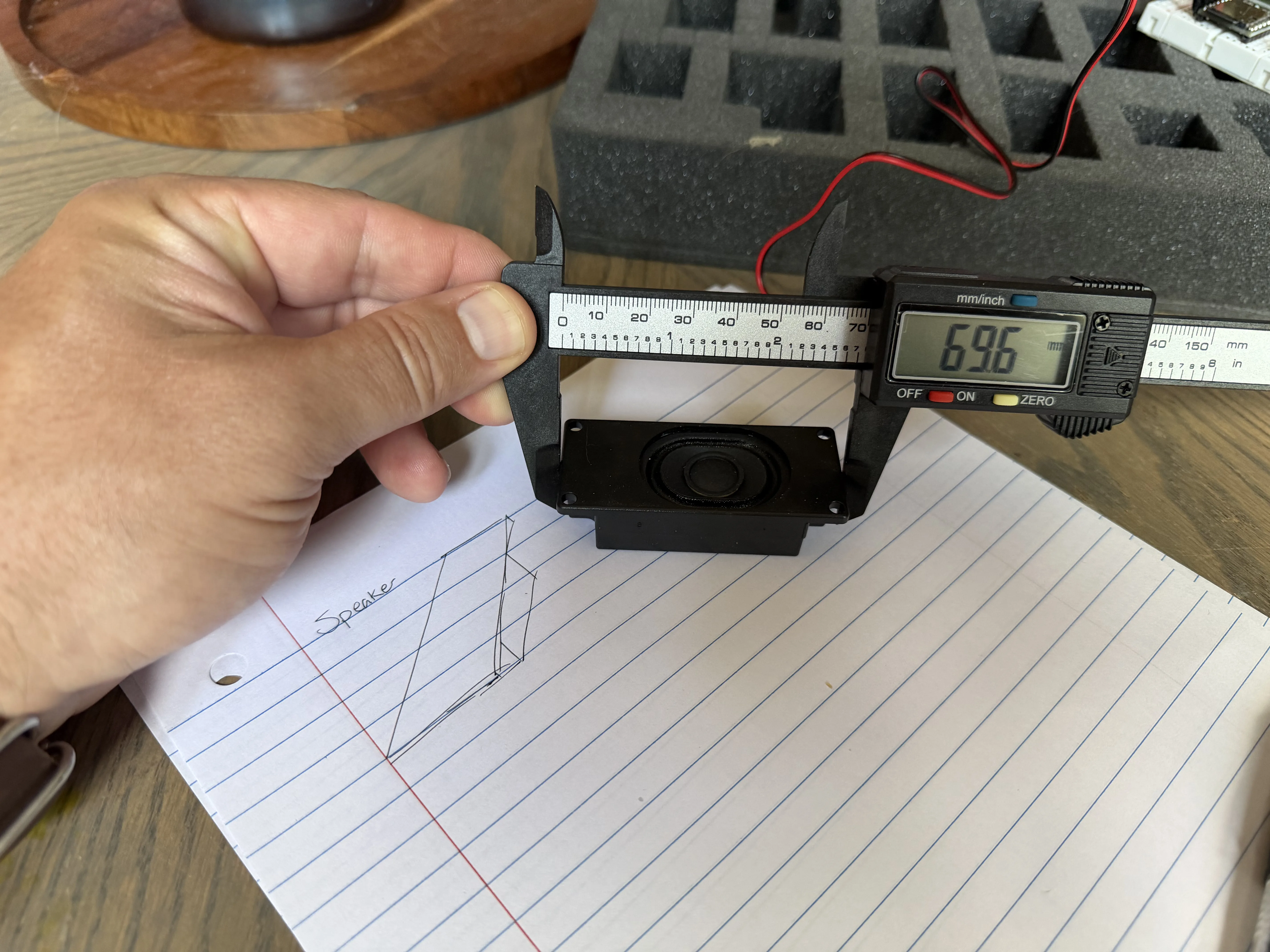 Digital calipers measuring the speaker enclosure at 69.6mm, resting on a notebook page with a hand-drawn speaker sketch